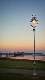Illuminated street light at riverbank against clear sky during sunset