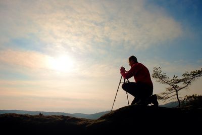 Nature photographer with tripod on cliff and thinking. dreamy fogy landscape, orange misty sunrise 