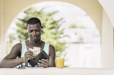 Content black male in earbuds taking bill out of wallet while sitting at table with glass of juice
