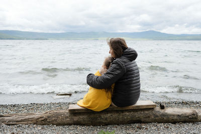 Back view of mother and daughter sitting hugging and kissing on the sea shore enjoying a winter day looking into distance