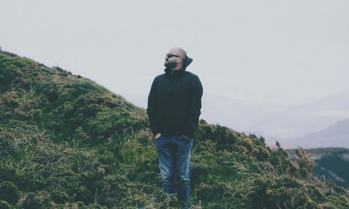 Man with hands in pockets standing on hill against sky