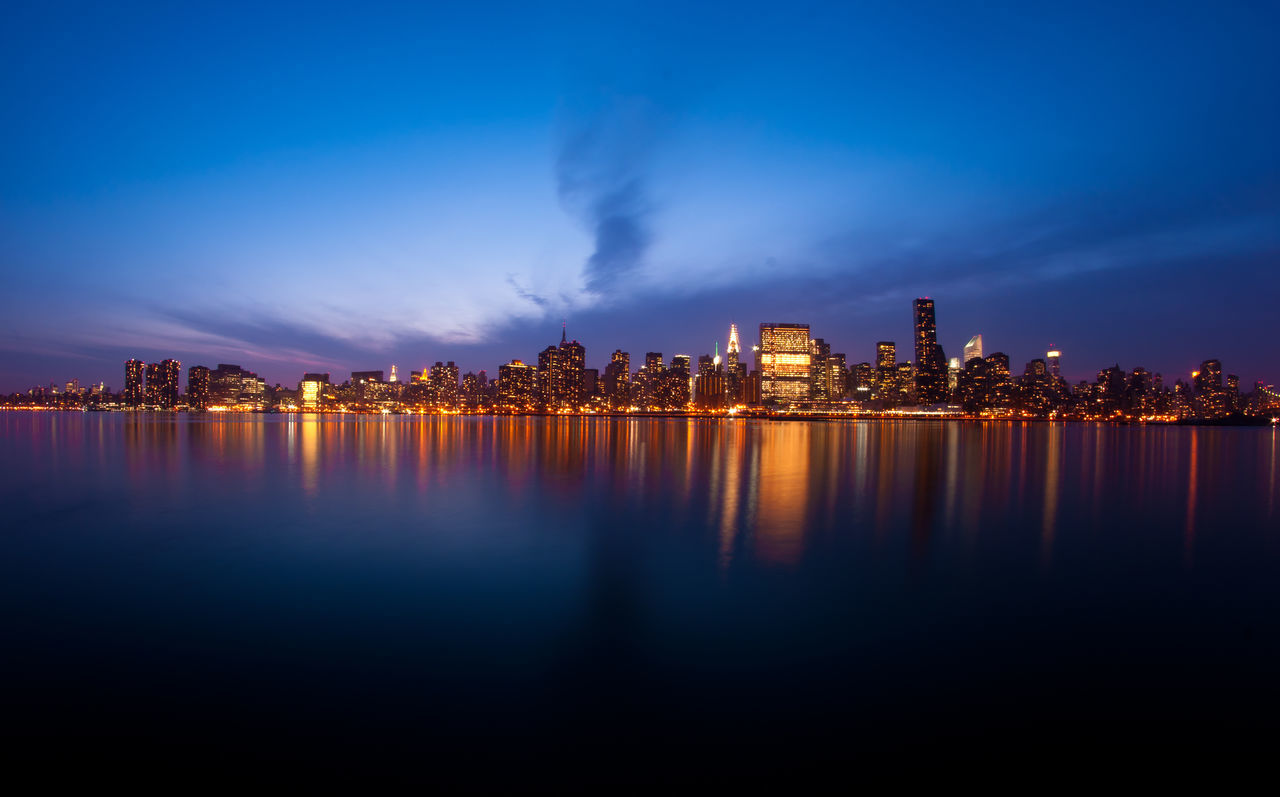 illuminated, water, city, building exterior, reflection, architecture, built structure, night, waterfront, sky, cityscape, river, skyscraper, blue, sea, urban skyline, dusk, mid distance, skyline, tall - high