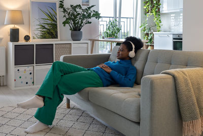 Happy relaxed african american woman in headphones closing eyes enjoying music sits on sofa