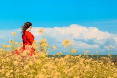 A woman in a bright red dress walks in the middle of a blooming yellow field on a clear summer day