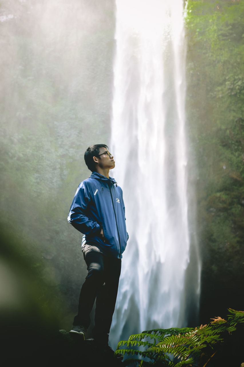 MAN STANDING BY WATERFALL AGAINST BLURRED BACKGROUND