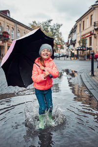 Happy smiling girl holding big umbrella jumping in the puddle during walk in a downtown on rainy day