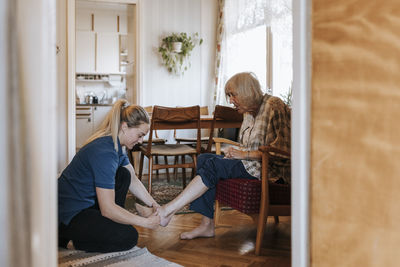 Side view of female care assistant helping senior woman wearing socks while sitting at home