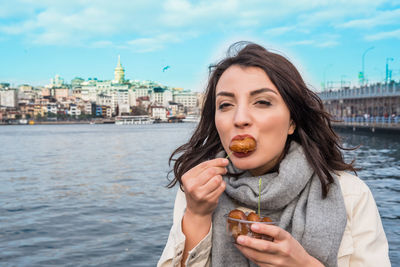 Portrait of woman eating food while standing against river in city