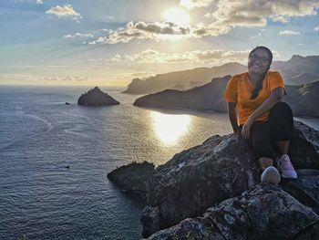 Smiling woman sitting on rock by sea against sky during sunset