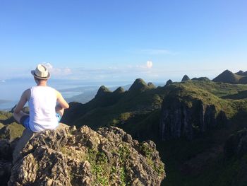 Rear view of man sitting on cliff by mountains against blue sky