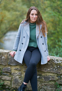 Portrait of a young caucasian woman with brown hair on a stone bridge with the green of the trees in the background. dressed in a gray jacket, green sweater, dark pants and military boots