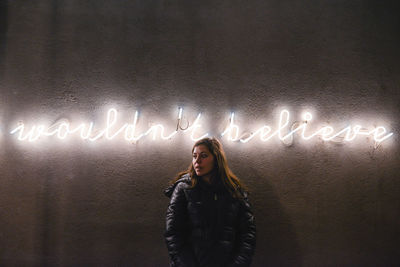 Woman looking away standing by illuminated text on wall