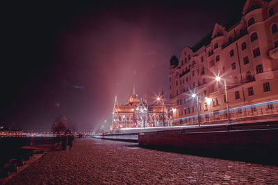 Illuminated street by buildings against sky at night