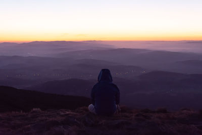 Rear view of woman sitting on landscape against sky at sunset