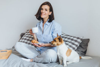 Woman with dog holding food and drink while sitting on bed against wall at home