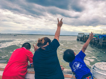 Rear view of woman with boys showing peace sign on pier in sea against cloudy sky