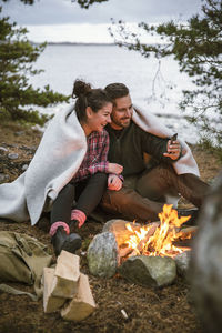 Couple wrapped in blanket using mobile phone while sitting by fire pit at campsite