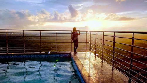 Woman standing by swimming pool against sky during sunset