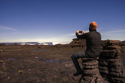 Rear view of male hiker photographing while sitting on rock against clear blue sky