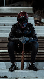 Full length of man in a leather jacket and red helmet sitting on bench and holding a baseball bat