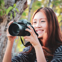 Close-up of woman smiling while photographing with camera against tree