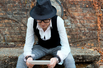 Mature man using digital tablet while sitting against old brick wall during autumn