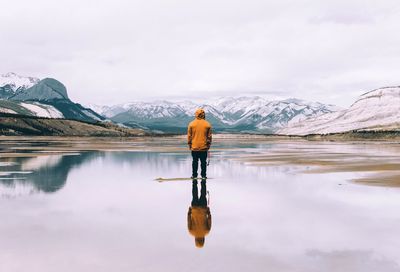Rear view of man standing amidst lake by snowcapped mountains
