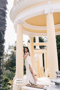 A beautiful brunette lady in an elegant wedding dress poses among the columns in the old city park