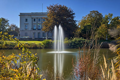 Fountain by lake against trees and building