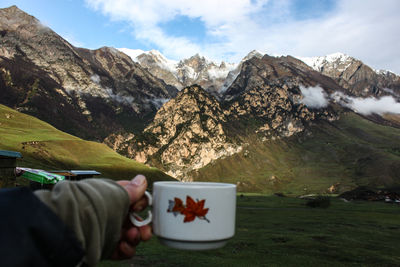 Cropped hand of person holding cup against snowcapped mountains