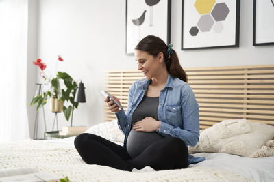 Pregnant woman using mobile phone while sitting on bed at home
