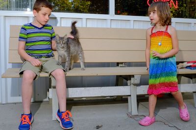 Girl looking at brother stroking cat while sitting on bench at park
