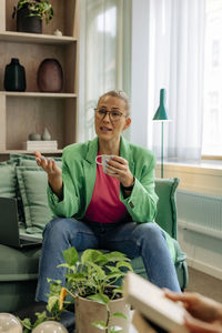 Mature female boss talking and holding cup while sitting on sofa at home