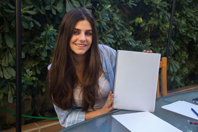 Portrait of smiling woman holding spiral notebook against plants