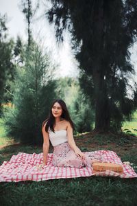 Young woman looking away while sitting on picnic blanket against trees