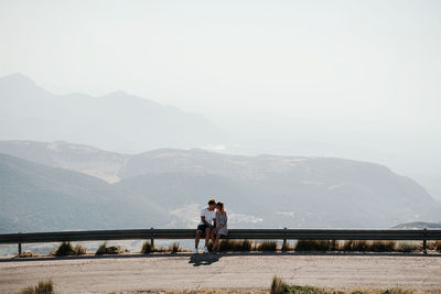 From below young man embracing and kissing girlfriend while sitting on rail of roadside against cloudless sky