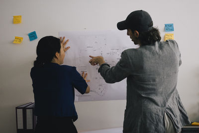 Rear view of multiracial architect colleagues discussing over blueprint on wall at home office