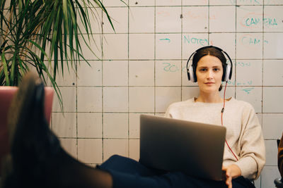 Portrait of confident young female computer hacker listening through headphones while sitting with laptop against tile w