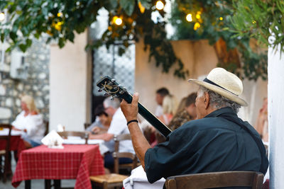 Rear view of man playing guitar while sitting on chair at outdoor restaurant