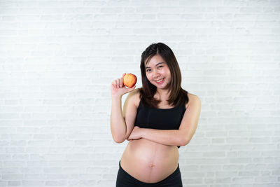 Portrait of smiling pregnant woman holding apple while standing against wall