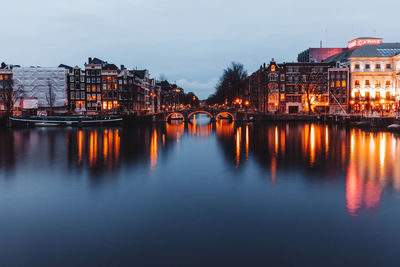 Illuminated bridge over river by buildings against sky at dusk