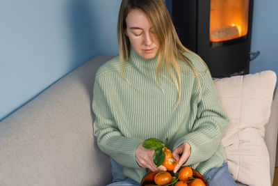 Blonde woman peels a tangerine while sitting on a sofa by the fireplace.