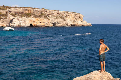 Side view of shirtless man standing on rock by sea against clear sky