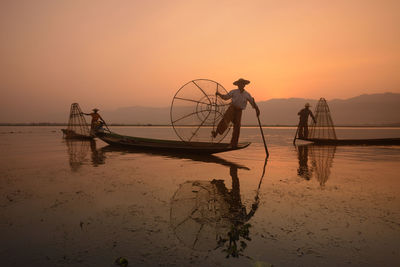 Fishermen fishing in lake against sky during sunset