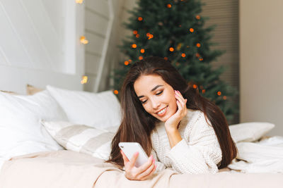 Young asian woman in cozy white knitted sweater using mobile on bed in the room with christmas tree