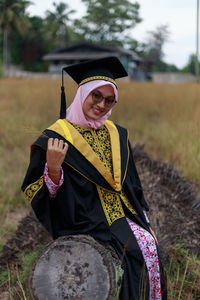 Portrait of woman in graduation gown sitting on log