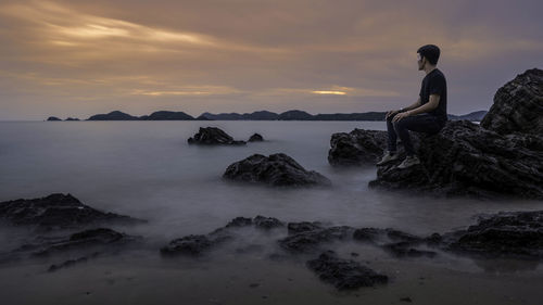 Man sitting on rock by sea against sky during sunset