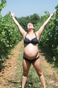 Pregnant woman with arms raised wearing bikini while standing on agricultural field during sunny day