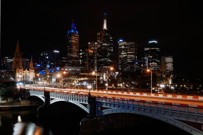 Illuminated bridge over river by buildings against sky at night