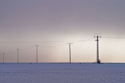 Mid distant view of electricity pylons on snow covered field against sky during sunset